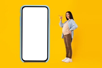 Cool app for pregnancy. Happy pregnant lady pointing at giant blank cellphone with empty screen, collage, mockup