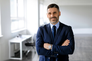 Portrait of confident middle aged male entrepreneur standing with folded arms and smiling at camera, posing in office