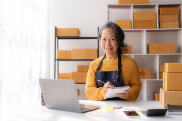 Asian senior female e-commerce entrepreneur checking goods for delivery to customer and using laptop.