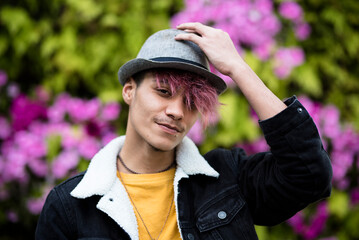 Portrait of cute nice handsome teenager alternative style looking on camera and smile touching hat on violet hair. SPring season park background flowers and green plants