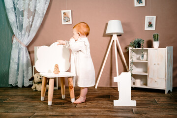 Little redhead baby girl celebrates first birthday anniversary. 1 year family party Professional photoshoot in photo studio. Cute adorable red hair kid in white vintage dress. Children's room at home