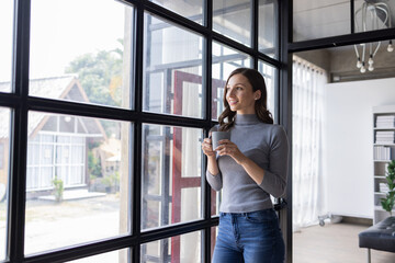 Portrait of young canadian female freelancer successful businesswoman holding cup of hot beverage tea coffee looking at the side thinking in workplace.