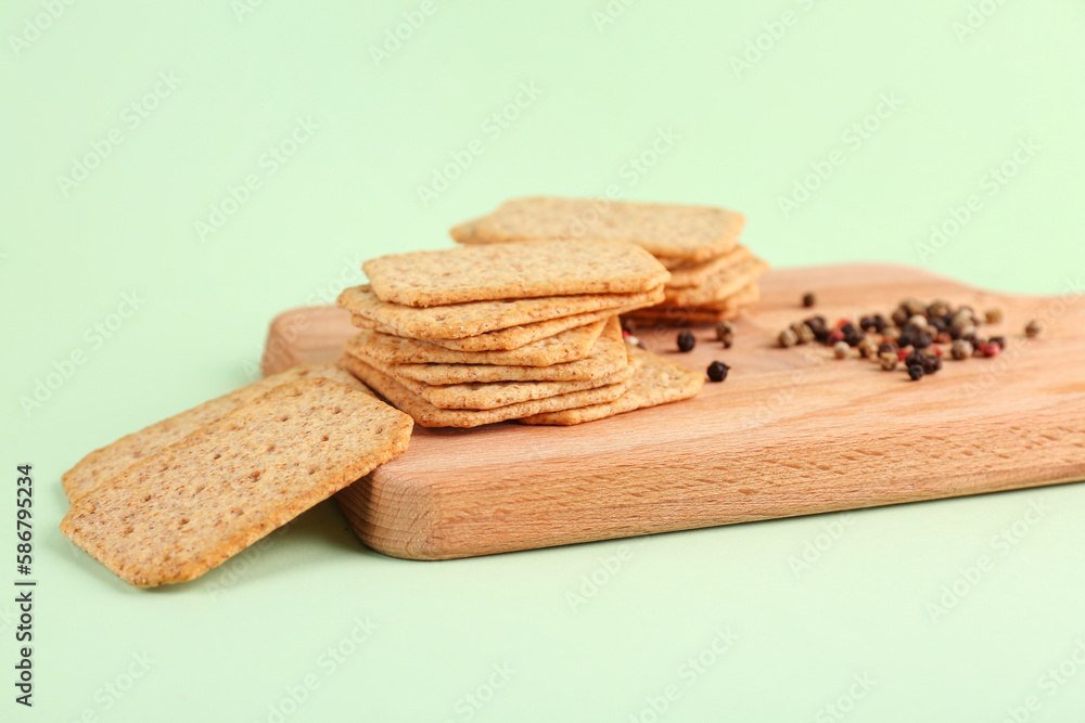 Board with tasty crackers and peppercorn on green background