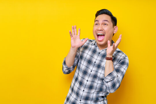 handsome young asian man surprised in plaid shirt looks aside and spreads his hands say wow isolated