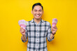 © Bangun Stock Photo - Portrait of cheerful young Asian man wearing plaid shirt Holding fan of cash money in rupiah banknotes isolated on yellow background. people lifestyle concept