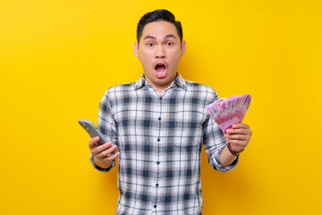 Wall Mural - Portrait of Surprised young Asian man wearing plaid shirt holding mobile phone and cash money in rupiah banknotes isolated on yellow background. people lifestyle concept