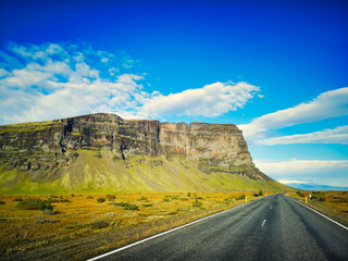 An open road in Iceland leads towards a massive cliff under a bright blue sky, surrounded by grassy plains and a scenic landscape