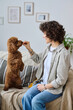 © AnnaStills - Vertical image of young woman sitting on sofa and training her little dog and giving her food