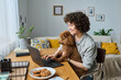 © AnnaStills - Young woman sitting at table with her dog and using laptop to do her online work