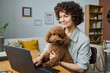 © AnnaStills - Young woman sitting at table and hugging her dog while working online on laptop in the room