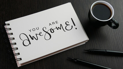 YOU ARE AWESOME! black lettering in notebook with cup of coffee and pens on black wooden desk