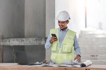 Engineer or Architect with protective safety helmet checking using smartphone holding architectural drawing at construction site. Engineering, Architecture and building project concepts