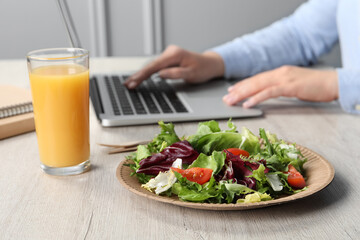 Office employee working with laptop at white wooden table, focus on fresh vegetable salad with glass of juice. Business lunch