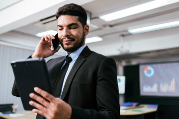 Smart and positive businessman call and talk on a smartphone alone while watching a digital tablet in the office. Professional making business calls and enjoying corporate mobile conversation indoors.