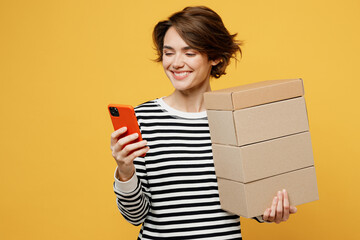 Young happy woman wear casual striped black and white shirt hold in hand stack cardboard blank boxes use mobile cell phone isolated on plain yellow color background studio portrait. Lifestyle concept.