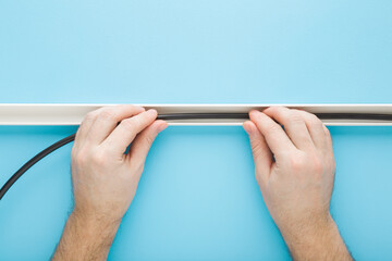 Wall Mural - Young adult man hands putting dark black new wire in white plastic channel on light blue wall background. Pastel color. Closeup. Point of view shot. Electrician working.