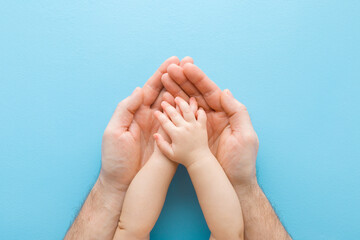 Wall Mural - Baby boy hands on young adult father palms on light blue table background. Pastel color. Closeup. Point of view shot. Lovely, emotional, sentimental moment. Safety concept. Top down view.