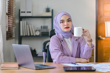 Wall Mural - Portrait of a beautiful Muslim millennial Asian businesswoman wearing a purple hijab working remotely from her home office. Looking at camera