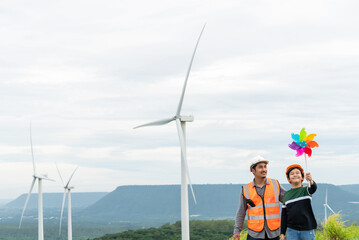 Wall Mural - Engineer with his son holding windmill toy on a wind farm atop a hill or mountain. Progressive ideal for the future production of renewable, sustainable energy. Energy generated from wind turbine.