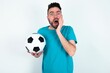 © Jihan - Stupefied Young man holding a ball over white background expresses excitement and thrill, keeps jaw dropped, hands on cheeks, has eyes popped out