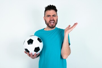 Wall Mural - Indignant Young man holding a ball over white background gestures in bewilderment, frowns face with dissatisfaction.