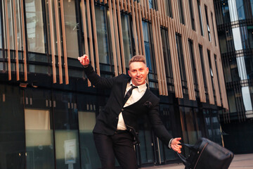 Happy young man businessman in suit with briefcase having fun at business building corporate company, looking at camera. Joyful guy near office house located in financial district. Copy ad text space