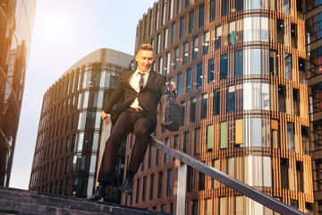 Happy young man businessman in suit with briefcase having fun near business building corporate company, slides down railing of stairs. Joyful guy near office house in financial district. Copy ad space