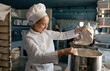 Female confectioner wearing white uniform putting flour into big metal bowl