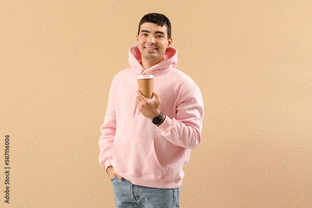 Stylish young man in hoodie drinking coffee on beige background