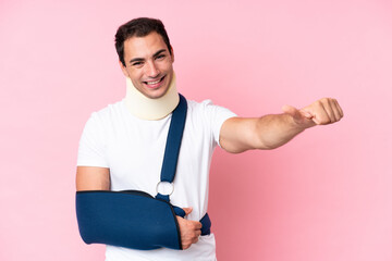 Wall Mural - Young caucasian man with broken arm and wearing a sling isolated on pink background giving a thumbs up gesture