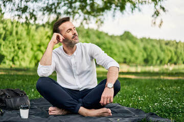 Middle-aged man sitting cross-legged on blanket in park daydreaming feeling relaxed