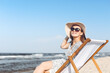 © rogerphoto - Happy brunette woman wearing sunglasses and hat relaxing on a wooden deck chair at the ocean beach