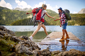 young couple on wooden jetty by lake. woman jumping, man lending a hand to her. summer trip in natur