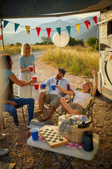 Group of friends in front of camper rv sitting and cheering with drinks. Roadtrip summer adventure. Fun, togetherness, travel, nature concept.