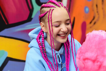 Caucasian teenage hipster girl with pink braids eats cotton candy against the background of a multicolored street wall.Summer concept.Generation Z style.