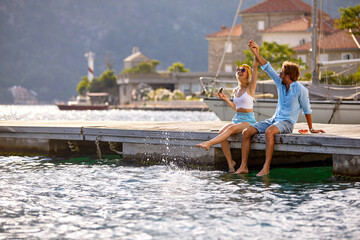 Wall Mural - Young cheerful couple holding hands up in air, enjoying and celebrating summer holiday on wooden jetty by water. Tourism, summertime, togetherness, lifestyle concept.