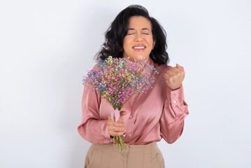 Wall Mural - young woman holds big bouquet of nice flowers over white background being excited for success with raised arms and closed eyes celebrating victory. Winner concept.