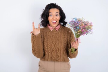 young woman holds big bouquet of nice flowers over white background makes rock n roll sign looks self confident and cheerful enjoys cool music at party. Body language concept.