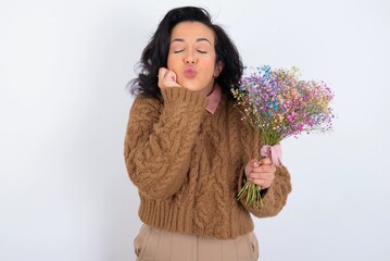 young woman holds big bouquet of nice flowers over white background with surprised expression keeps hands under chin keeps lips folded makes funny grimace