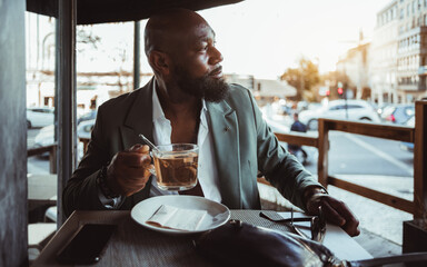 A portrait of a bald, black man with a beard at a coffee place by the side of a street. Dressed in a stylish suit jacket and white shirt, holds a cup of tea, exuding a sense of calm and sophistication