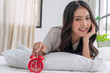 © kaew6566 - Beautiful cheerful asian woman early wake up lying relax on pillow holding hand red alarm clock at home. Happy young girl refreshing early wake up on white with alarm clock looking camera in morning.