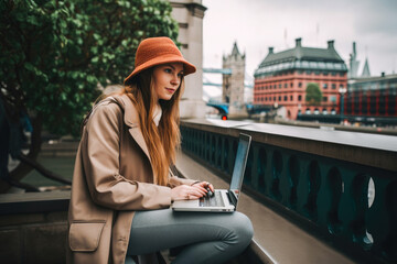 Wall Mural - Young woman sitting in London and doing work on laptop. Student or freelancer lifestyle. Tower Bridge in the background. High quality generative ai