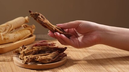 Canvas Print - A moment woman hand holding red ginseng root and gently shaking it on blurred background with brown mosaic and ginseng roots placed on wooden plate. Herbs popular at China and Korea