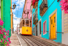 Lisbon, Portugal - Yellow Tram On A Street With Colorful Houses And Flowers On The Balconies - Bica Elevator Going Down The Hill Of Chiado.