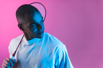 Racket sport concept. Stunning indoor portrait of young adult male athlete with badminton racket behind his neck looking aside. Pink background and contrasting blue light. High quality photo