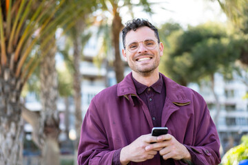 Wall Mural - Young caucasian man at outdoors looking at the camera and smiling while using the mobile