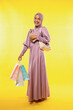 © Odua Images - Asian woman wearing color dress with hijab carrying shopping bags and mobile phone while shopping on yellow background