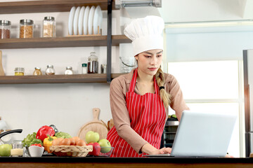 Happy smiling beautiful Asian woman wear apron chef hat at kitchen, look at laptop computer for searching recipes online, use internet for cooking tasty meal, female chef cooks delicious healthy food.