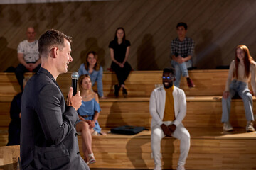 Young caucasian male speaker giving talk with microphone in conference hall at business meeting event. Group of people in audience at conference hall, listen to lesson. Business and entrepreneurship