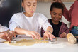 © Ladanifer - Close up of cousins cutting cookie shapes in a cookie dough in the kitchen with their family.